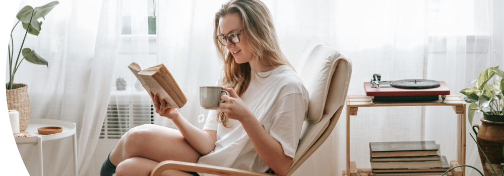 Frau sitzt mit Buch und Kaffee lesend im Sessel